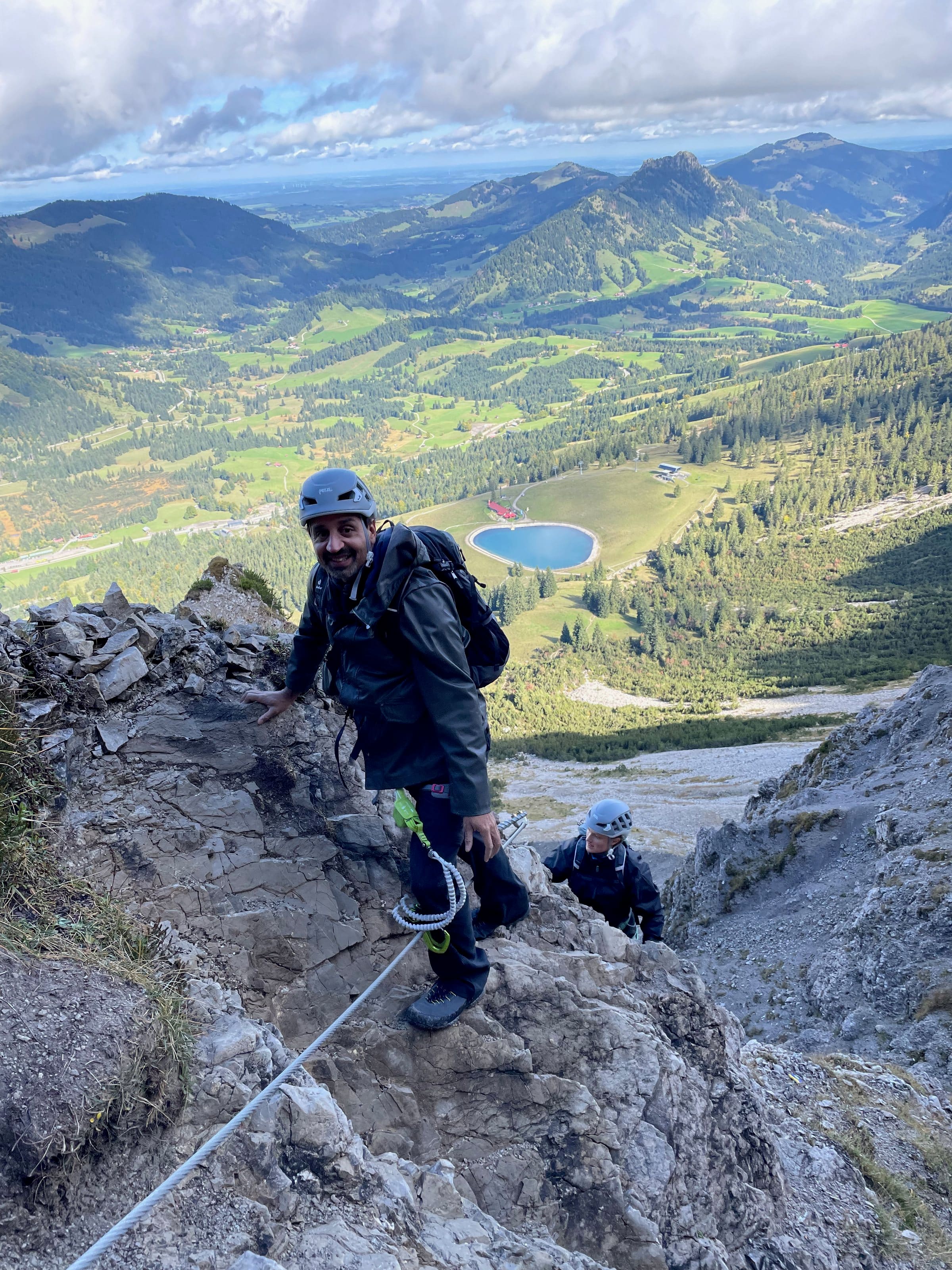 Dave mit Helm am Klettersteig, unter ihm ein Tal mit See